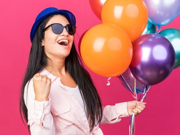 Excited young beautiful girl wearing party hat and glasses holding balloons showing yes gesture 