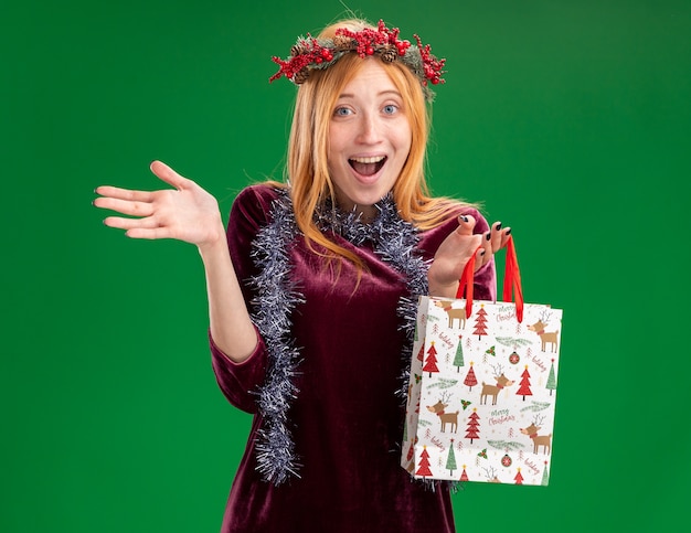 Бесплатное фото Возбужденная молодая красивая девушка в красном платье с венком и гирляндой на шее держит подарочный пакет, протягивая руку, изолированную на зеленой стене