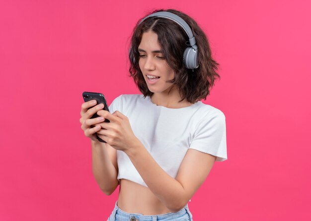 Возбужденная молодая красивая женщина в наушниках и держащая мобильный телефон на розовой стене с копией пространства