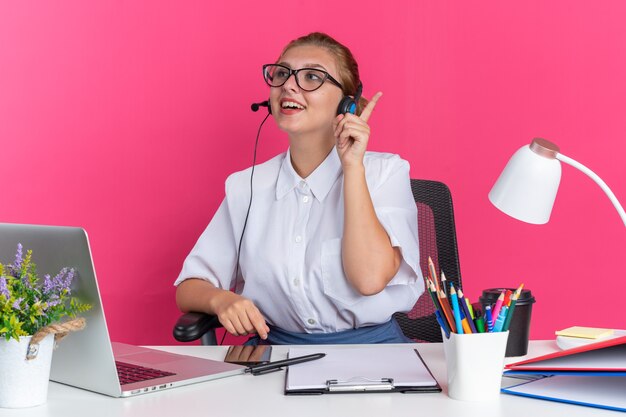 Excited young blonde call centre girl wearing headset and glasses sitting at desk with work tools looking at side pointing up isolated on pink wall