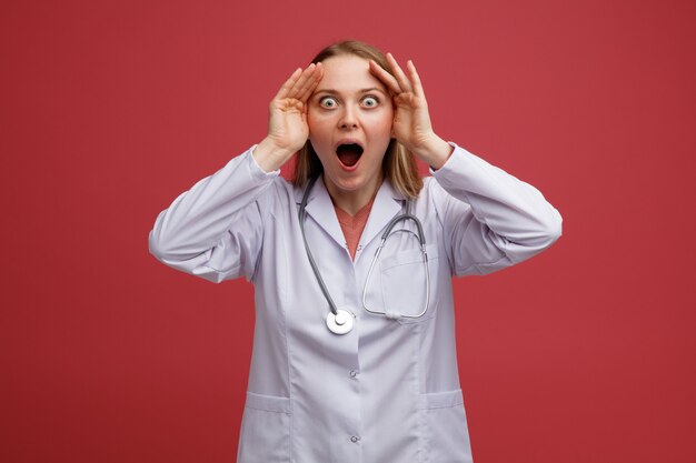 Excited young blonde female doctor wearing medical robe and stethoscope around neck keeping hands on head 