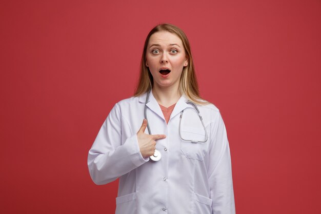 Возбужденная молодая блондинка женщина-врач в медицинском халате и стетоскопе на шее, указывая на себя