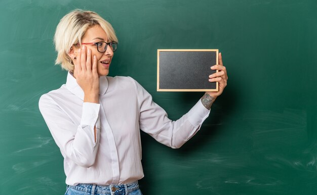 excited young blonde female teacher wearing glasses in classroom standing in front of chalkboard showing mini blackboard looking at it keeping hand on face with copy space