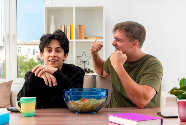 Excited young blonde handsome guy keeping fists up sitting at table and looking at smiling young brunette handsome guy holding hands together and looking 