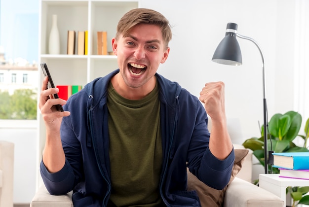 Free photo excited young blonde handsome man sits on armchair keeping fist and holding phone