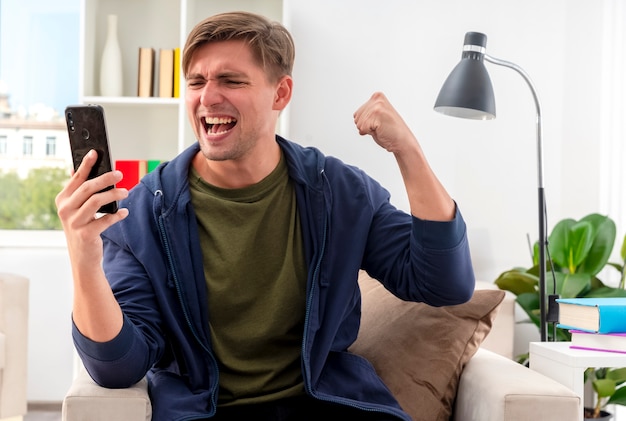 Excited young blonde handsome man sits on armchair keeping fist and looking at phone