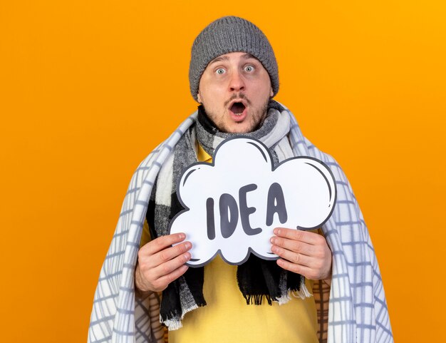 Excited young blonde ill slavic man wearing winter hat and scarf wrapped in plaid holds idea bubble isolated on orange wall with copy space