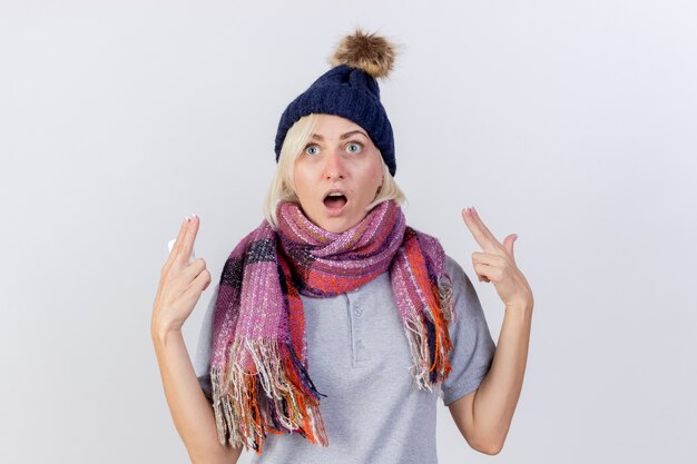 Excited young blonde ill slavic woman wearing winter hat and scarf points up with two hands holding pack of medical pills isolated on white wall with copy space