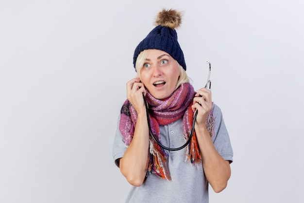 Бесплатное фото Возбужденная молодая блондинка больная женщина в зимней шапке и шарфе держит стетоскоп на белой стене