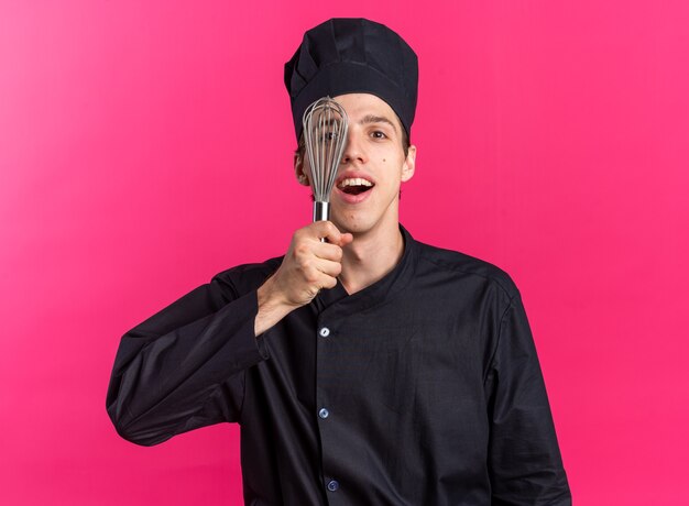 Excited young blonde male cook in chef uniform and cap looking at camera covering half of face with whisk isolated on pink wall