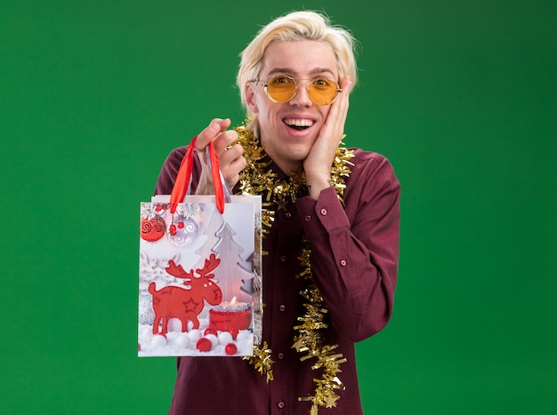 Eccitato giovane uomo biondo con gli occhiali con la ghirlanda di orpelli intorno al collo che tiene il sacchetto del regalo di natale mantenendo la mano sul viso isolato sulla parete verde con lo spazio della copia