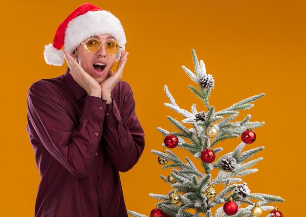 오렌지 벽에 고립 된 얼굴 근처에 손을 유지 장식 된 크리스마스 트리 근처에 서 산타 모자와 안경을 쓰고 흥분된 젊은 금발의 남자