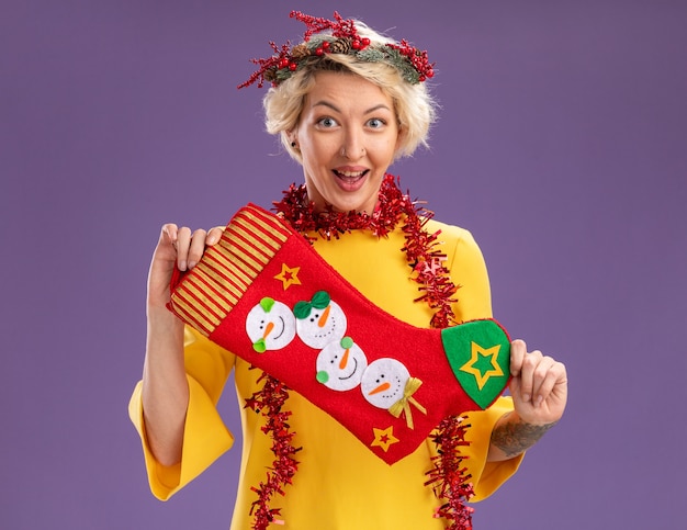 Бесплатное фото Возбужденная молодая блондинка в рождественском венке и гирлянде из мишуры на шее, держащая рождественский чулок, изолирована на фиолетовой стене