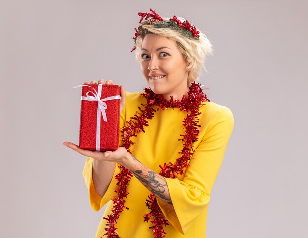 Возбужденная молодая блондинка в рождественском венке и гирлянде из мишуры на шее, держащая рождественский подарочный пакет, прикусывает губу, изолированную на белой стене с копией пространства