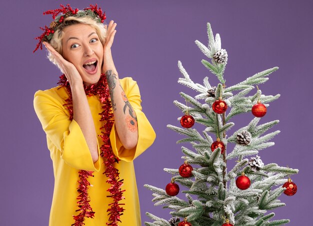 Excited young blonde woman wearing christmas head wreath and tinsel garland around neck standing near decorated christmas tree keeping hands on face looking  isolated on purple wall