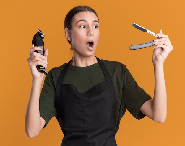 Foto gratuita eccitata giovane barbiere bruna in uniforme tiene in mano un tagliacapelli e guarda un rasoio dritto