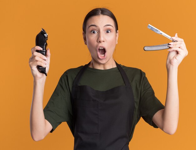 Excited young brunette barber girl in uniform holds hair clippers and straight razor