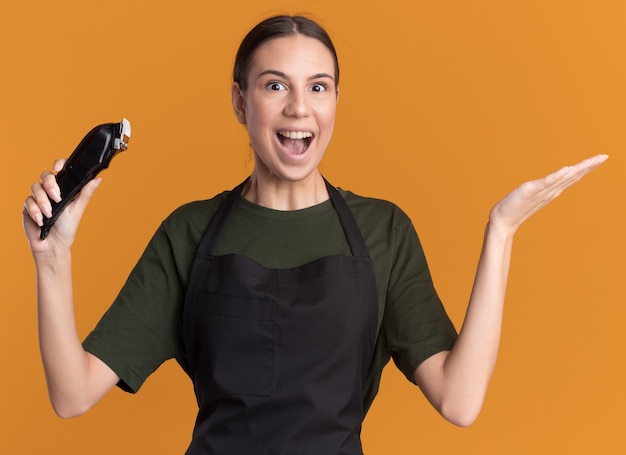 Excited young brunette barber girl in uniform keeps hand open and holds hair clippers isolated on orange wall with copy space