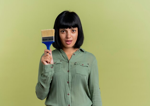 Excited young brunette caucasian girl holds paint brush and looks at camera isolated on olive green background with copy space