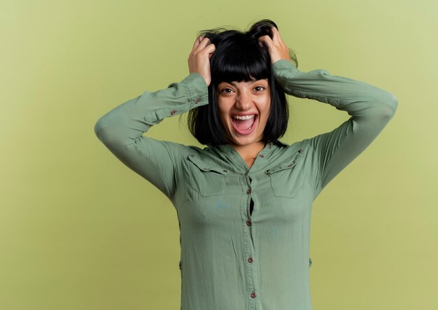 Free photo excited young brunette caucasian girl lift hair looking at camera isolated on olive green background with copy space