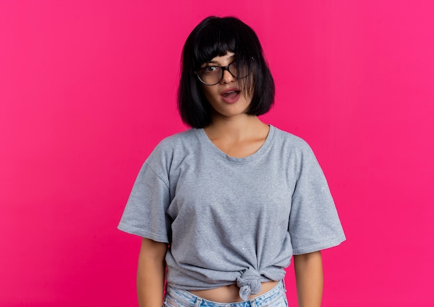 Excited young brunette caucasian girl in optical glasses looks at camera isolated on pink background with copy space