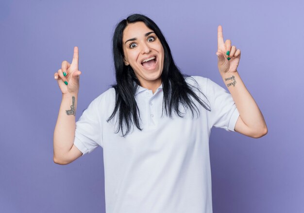 Excited young brunette caucasian girl points up and looks isolated on purple wall