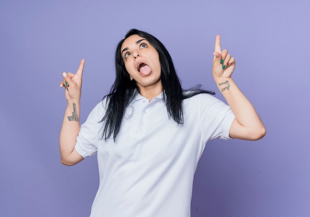 Excited young brunette caucasian girl sticks out tongue and points up isolated on purple wall