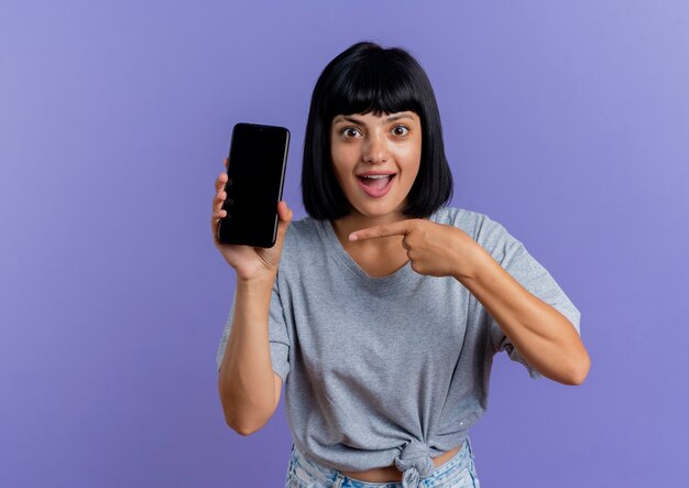 Excited young brunette caucasian woman holds and points at phone