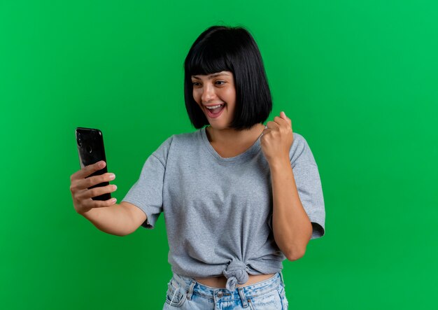 Excited young brunette caucasian woman keeps fist and looks at phone isolated on green background with copy space