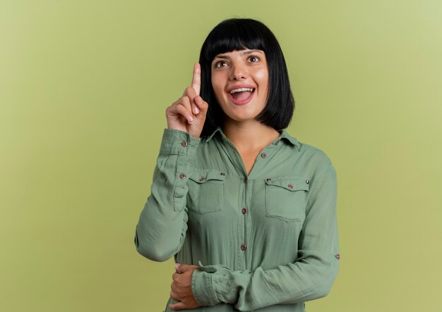 Excited young brunette caucasian woman points up isolated on olive green background with copy space