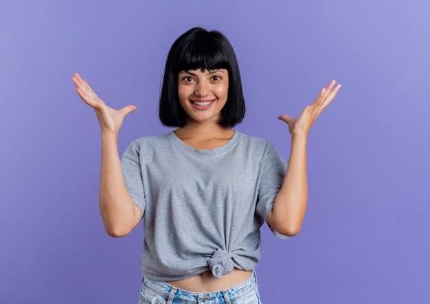 Excited young brunette caucasian woman stands with raised hands looking at camera isolated on purple background with copy space