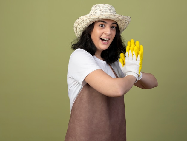 Возбужденная молодая брюнетка женщина-садовник в униформе в садовой шляпе и перчатках держит руки вместе, изолированные на оливково-зеленой стене