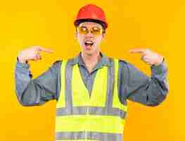 Free photo excited young builder man in uniform wearing glasses points at himself