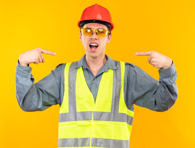 Возбужденный молодой строитель в униформе в очках указывает на себя
