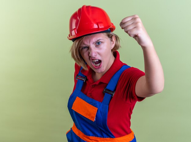 возбужденная молодая женщина-строитель в униформе, показывающая жест `` да '' на оливково-зеленой стене