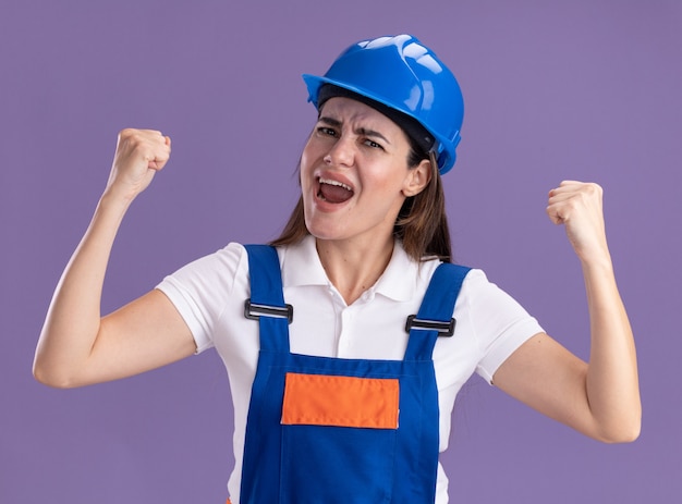 Free photo excited young builder woman in uniform showing yes gesture isolated on purple wall