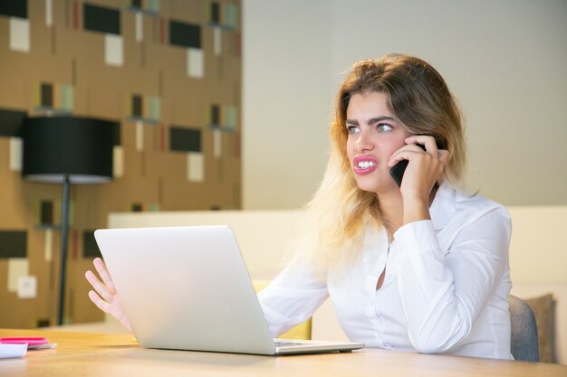 Возбужденная молодая деловая женщина разговаривает по мобильному телефону в коворкинге, сидя за столом с ноутбуком