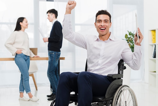 Excited young businessman holding digital tablet in hand sitting on wheelchair with business couple looking at each other
