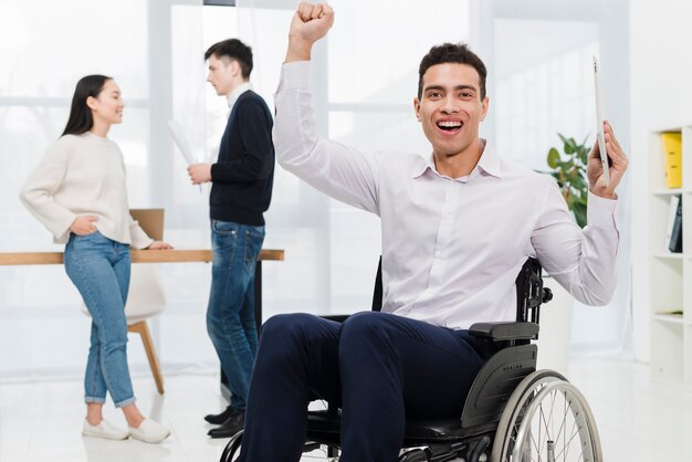 Возбужденный молодой бизнесмен, держа в руке цифровой планшет, сидя на инвалидной коляске с парами бизнес, глядя друг на друга