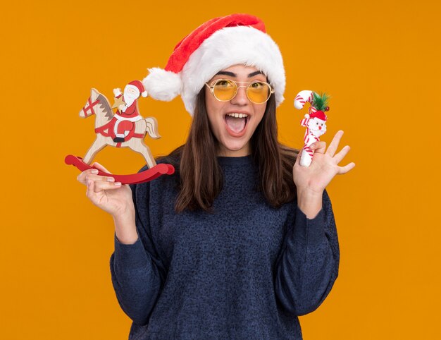 Excited young caucasian girl in sun glasses with santa hat holds santa on rocking horse decoration and candy cane isolated on orange wall with copy space