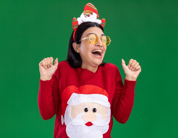 Excited young caucasian girl wearing santa claus headband and sweater with glasses looking at camera showing thumbs up isolated on green background