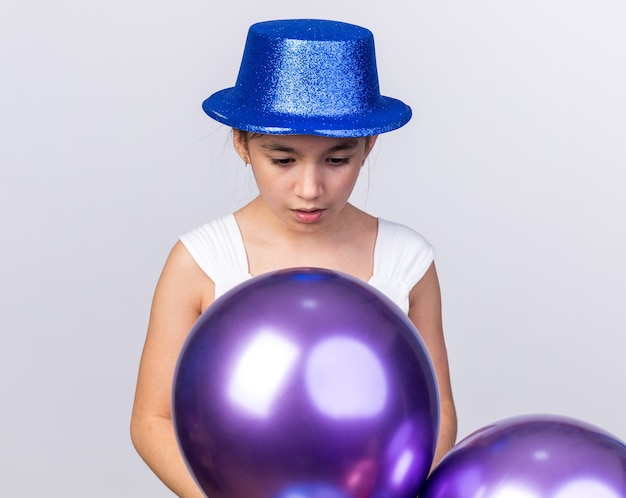 Foto gratuita eccitata giovane ragazza caucasica con cappello blu partito guardando palloncini di elio isolati sulla parete bianca con lo spazio della copia