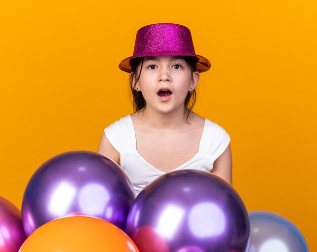 excited young caucasian girl with purple party hat standing with helium balloons  isolated on orange wall with copy space