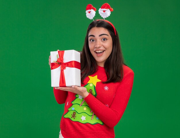 excited young caucasian girl with santa headband holds christmas gift box isolated on green background with copy space