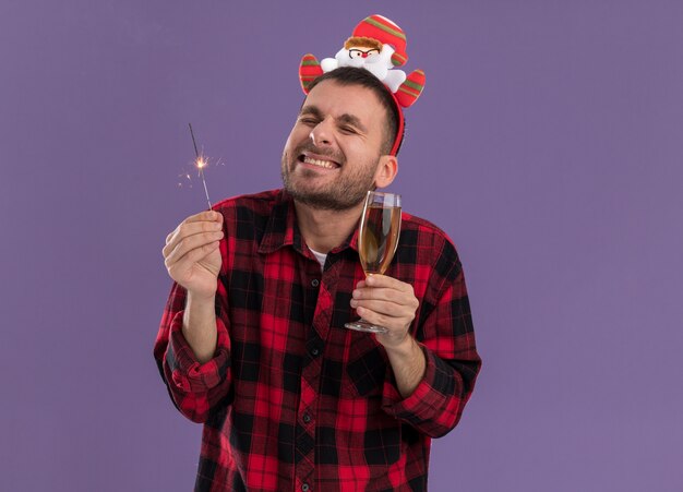 Excited young caucasian man wearing santa claus headband holding holiday sparkler and glass of champagne smiling with closed eyes isolated on purple wall with copy space