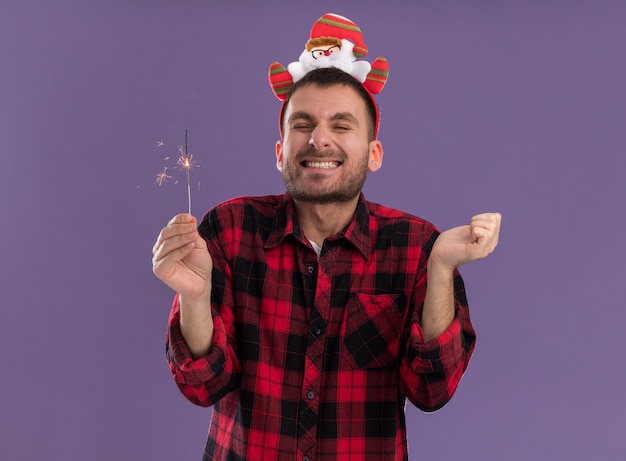 Excited young caucasian man wearing santa claus headband holding holiday sparkler keeping hand in air smiling with closed eyes isolated on purple wall