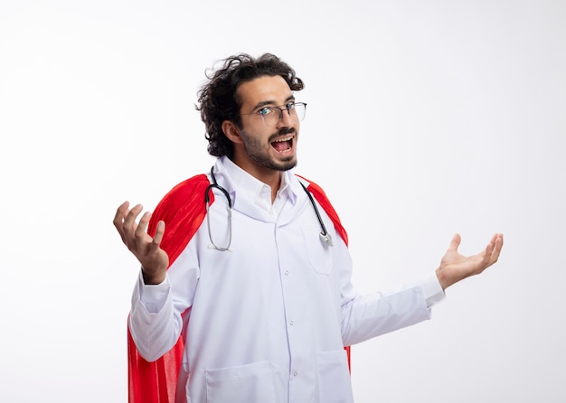 Excited young caucasian superhero man in optical glasses wearing doctor uniform with red cloak and with stethoscope around neck holds hands open 