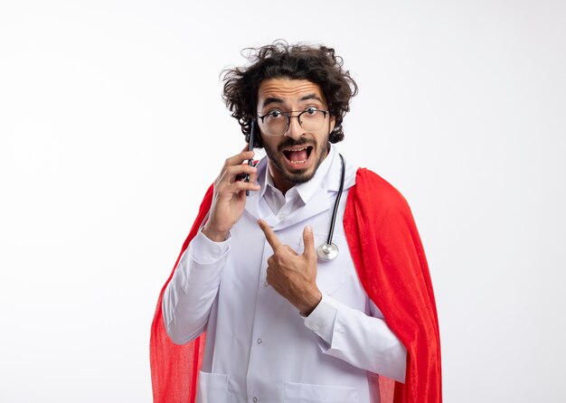 Excited young caucasian superhero man in optical glasses wearing doctor uniform with red cloak and with stethoscope around neck pointing and talking on phone isolated on white wall