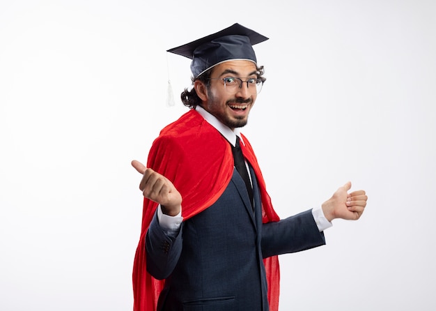 Excited young caucasian superhero man in optical glasses wearing suit with red cloak and graduation cap stands sideways pointing at sides 