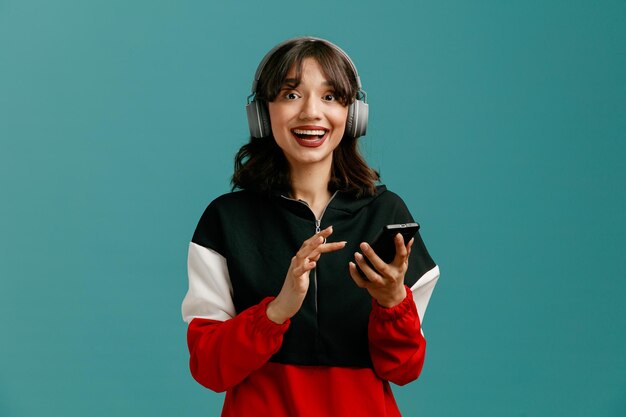 Возбужденная молодая кавказская женщина в наушниках держит мобильный телефон и смотрит в камеру на синем фоне